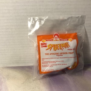Puede incluir: Una bolsa de pl&aacute;stico que contiene una figura de acci&oacute;n de Spiderman roja y azul. La bolsa es naranja y tiene las palabras "Marvel Comics" y "Spiderman" impresas en ella. El texto "The Amazing Spiderman Figurine" tambi&eacute;n est&aacute; impreso en la bolsa.