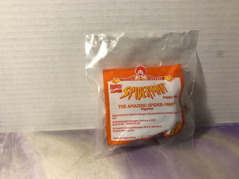 Puede incluir: Una bolsa de pl&aacute;stico que contiene una figura de acci&oacute;n de Spiderman roja y azul. La bolsa es naranja y tiene las palabras "Marvel Comics" y "Spiderman" impresas en ella. El texto "The Amazing Spiderman Figurine" tambi&eacute;n est&aacute; impreso en la bolsa.