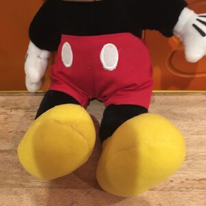 Puede incluir: Un juguete de peluche de Mickey Mouse sentado con las piernas cruzadas. Lleva pantalones cortos rojos con botones blancos, zapatos negros y guantes blancos. Tiene una cara blanca y negra con una nariz roja y una gran sonrisa.