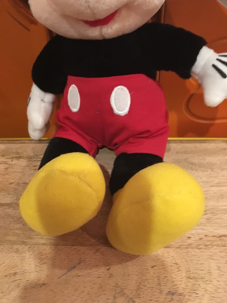 Puede incluir: Un juguete de peluche de Mickey Mouse sentado con las piernas cruzadas. Lleva pantalones cortos rojos con botones blancos, zapatos negros y guantes blancos. Tiene una cara blanca y negra con una nariz roja y una gran sonrisa.