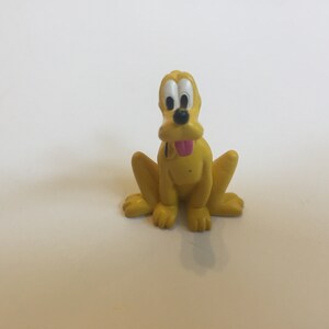 Puede incluir: Una figurita de plástico amarilla de Pluto, un perro de dibujos animados de Disney. Pluto está sentado con la lengua fuera y tiene ojos negros y nariz negra.