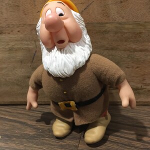 May include: A plush toy of the character Doc from the Disney film Snow White and the Seven Dwarfs. The toy is brown and yellow with a white beard and a surprised expression.