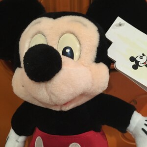 Puede incluir: Un juguete de peluche de Mickey Mouse con orejas negras, una nariz negra y un atuendo rojo y blanco. El juguete tiene una etiqueta blanca adherida a su oreja derecha.