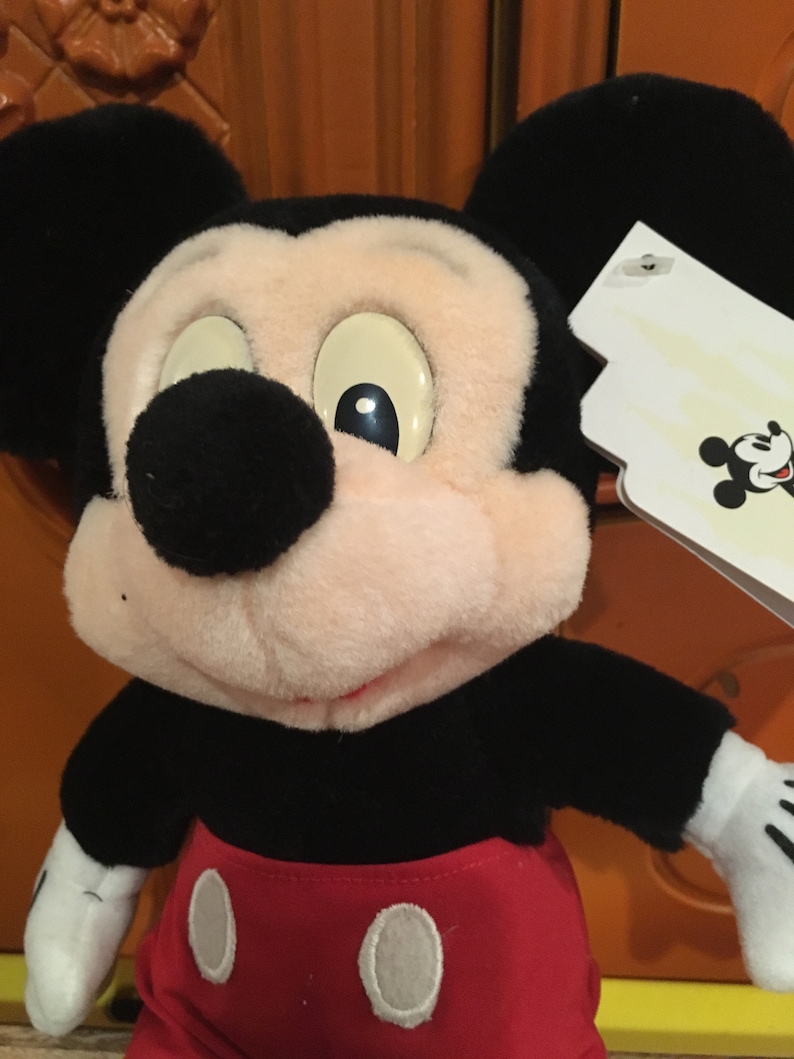 Puede incluir: Un juguete de peluche de Mickey Mouse con orejas negras, una nariz negra y un atuendo rojo y blanco. El juguete tiene una etiqueta blanca adherida a su oreja derecha.
