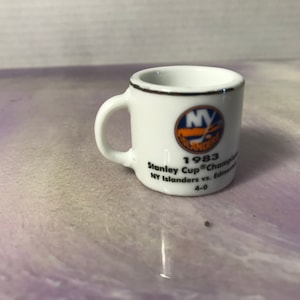 Może przedstawiać: Biały kubek ceramiczny z niebiesko-pomarańczowym logo New York Islanders. Kubek ma napis "1983 Stanley Cup Champions NY Islanders vs. Edmonton 4-0".