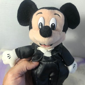 May include: A plush Mickey Mouse wearing a black tuxedo with a white bow tie. The plush toy has a black nose, large black ears, and a white face.