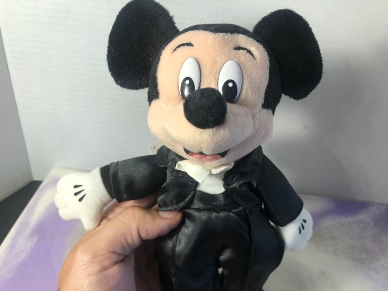 May include: A plush Mickey Mouse wearing a black tuxedo with a white bow tie. The plush toy has a black nose, large black ears, and a white face.
