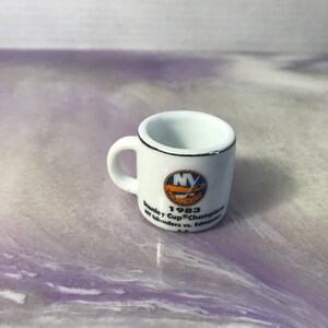 Może przedstawiać: Biały kubek ceramiczny z niebiesko-pomarańczowym logo New York Islanders. Kubek upamiętnia mistrzostwo Pucharu Stanleya z 1983 roku z tekstem "1983 Stanley Cup® Champions NY Islanders vs. Edmonton 4-0."