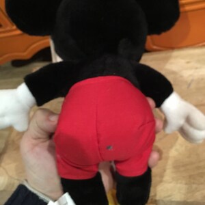 Może przedstawiać: Pluszowa zabawka Mickey Mouse w kolorze czarnym i czerwonym z ż&oacute;łtymi butami. Zabawka jest trzymana przez rękę.