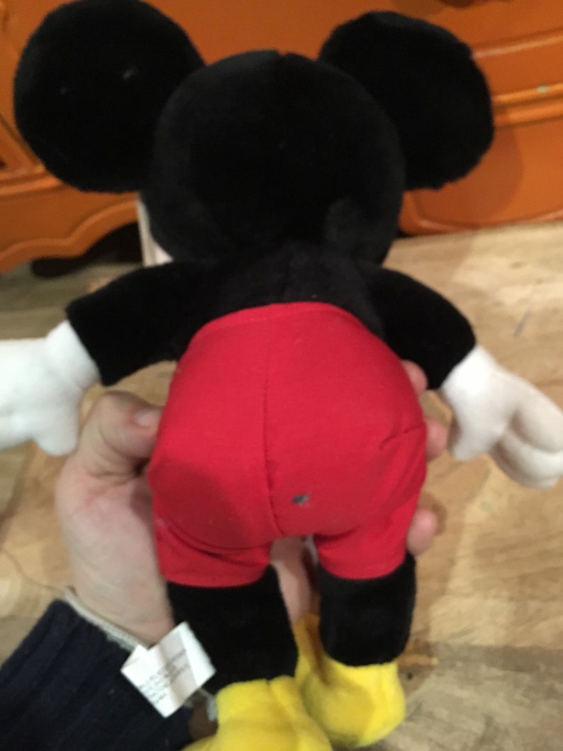 Może przedstawiać: Pluszowa zabawka Mickey Mouse w kolorze czarnym i czerwonym z ż&oacute;łtymi butami. Zabawka jest trzymana przez rękę.