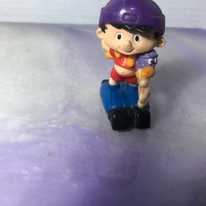 May include: A purple and blue figurine of a cartoon character riding a scooter. The character is wearing a purple hat and a red shirt. The character is holding a small purple and orange figurine.