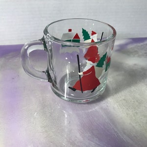 May include: A clear glass mug with a Christmas scene featuring a red-suited figure carrying a staff and a bag. The scene is painted in white, red, and green.