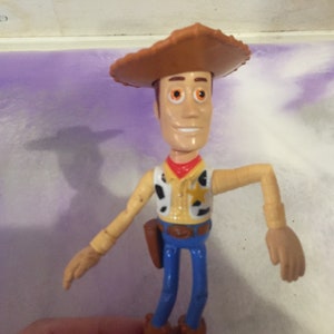 May include: A plastic toy figure of Woody from the Toy Story franchise. Woody is wearing his signature brown cowboy hat, a yellow vest with a sheriff's star, and blue jeans. He has a friendly smile and brown eyes.