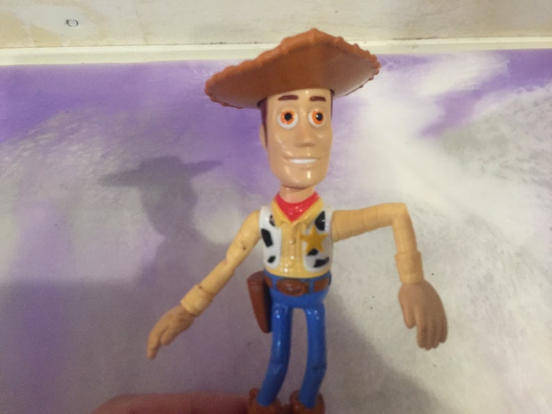 May include: A plastic toy figure of Woody from the Toy Story franchise. Woody is wearing his signature brown cowboy hat, a yellow vest with a sheriff's star, and blue jeans. He has a friendly smile and brown eyes.