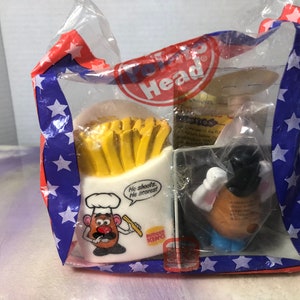 Pode incluir: Um saco de plástico contendo três brinquedos Mr. Potato Head. Um brinquedo é um chef com um chapéu do Burger King e uma caixa de batatas fritas. Outro brinquedo é um cowboy com um chapéu e uma arma. O terceiro brinquedo é um Mr. Potato Head com um chapéu-coco e um bigode. O saco tem um padrão de bandeira americana vermelha, branca e azul.