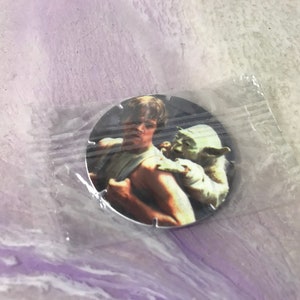 Può includere: Uno spillo rotondo color argento con un'immagine di Luke Skywalker e Yoda di Star Wars. Luke tiene Yoda tra le braccia. Lo sfondo è grigio chiaro.