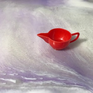 Può includere: Un piccolo e luminoso brocchetto di plastica rosso con manico.