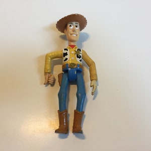 Op de afbeelding: Een plastic speelgoedfiguur van Woody uit de Toy Story-franchise. Woody draagt zijn kenmerkende bruine cowboyhoed, een geel vest met een sheriffster, een blauwe spijkerbroek en bruine laarzen. Hij heeft een vriendelijke uitdrukking op zijn gezicht.