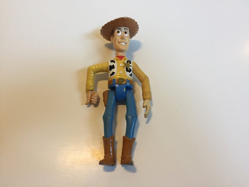 Op de afbeelding: Een plastic speelgoedfiguur van Woody uit de Toy Story-franchise. Woody draagt zijn kenmerkende bruine cowboyhoed, een geel vest met een sheriffster, een blauwe spijkerbroek en bruine laarzen. Hij heeft een vriendelijke uitdrukking op zijn gezicht.