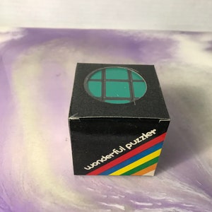 Może przedstawiać: Czarne pudełko z zielono-czarną kostką do układania puzzli z przodu. Na pudełku widnieje napis "wonderful puzzler" wydrukowany w kolorach tęczy.