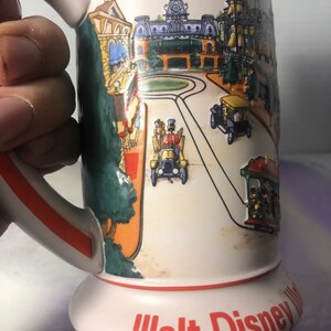 K&ouml;nnte beinhalten: Eine wei&szlig;e Keramiktasse mit rotem Rand und Henkel. Die Tasse zeigt eine farbenfrohe Illustration der Main Street, U.S.A. von Walt Disney World mit dem Text "Walt Disney World" in Rot am Boden.