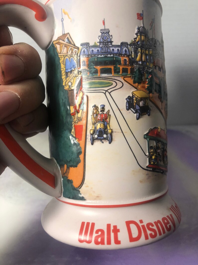 K&ouml;nnte beinhalten: Eine wei&szlig;e Keramiktasse mit rotem Rand und Henkel. Die Tasse zeigt eine farbenfrohe Illustration der Main Street, U.S.A. von Walt Disney World mit dem Text "Walt Disney World" in Rot am Boden.
