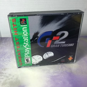 May include: A PlayStation Greatest Hits game case for Gran Turismo 2, a racing game. The case is black with a green PlayStation logo and a red, white, and blue G2 logo. The game is rated E for Everyone by the ESRB.