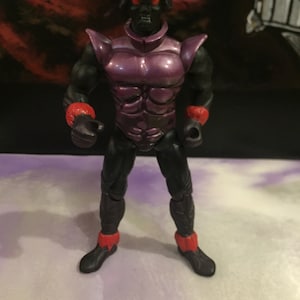 May include: A black and purple action figure with red accents. The figure has a bug-like head with red eyes and a black body with a purple chest piece. The figure is standing on a white surface.