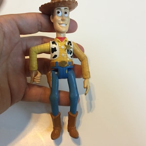 Op de afbeelding: Een plastic speelgoedfiguur van Woody uit de Toy Story-franchise. Woody draagt zijn kenmerkende bruine cowboyhoed, geel shirt, blauwe jeans en bruine laarzen. Hij heeft een vriendelijke uitdrukking op zijn gezicht.