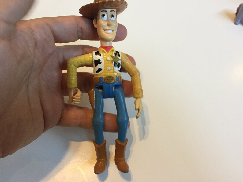 Op de afbeelding: Een plastic speelgoedfiguur van Woody uit de Toy Story-franchise. Woody draagt zijn kenmerkende bruine cowboyhoed, geel shirt, blauwe jeans en bruine laarzen. Hij heeft een vriendelijke uitdrukking op zijn gezicht.