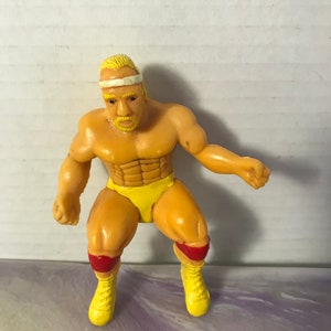 May include: A yellow plastic action figure of a wrestler with a white headband, yellow trunks, and red knee pads. The figure has a blonde wig and a beard.