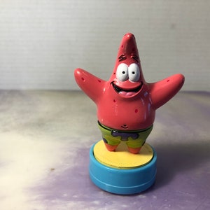 May include: A pink cartoon starfish character with a big smile and large eyes, wearing green shorts with a purple belt. The character is standing on a blue and yellow base.