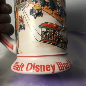 K&ouml;nnte beinhalten: Eine wei&szlig;e Keramiktasse mit einer bunten Illustration einer Stra&szlig;enszene in Walt Disney World. Die Tasse zeigt eine rote, gr&uuml;ne und gelbe Stra&szlig;enbahn mit Mickey Mouse und seinen Freunden, die darin fahren. Die Tasse hat einen roten Rand und die Worte "Walt Disney World" in roten Buchstaben auf dem Boden.