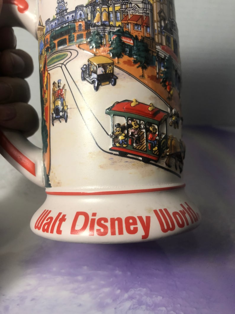 K&ouml;nnte beinhalten: Eine wei&szlig;e Keramiktasse mit einer bunten Illustration einer Stra&szlig;enszene in Walt Disney World. Die Tasse zeigt eine rote, gr&uuml;ne und gelbe Stra&szlig;enbahn mit Mickey Mouse und seinen Freunden, die darin fahren. Die Tasse hat einen roten Rand und die Worte "Walt Disney World" in roten Buchstaben auf dem Boden.