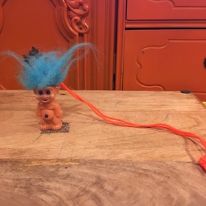 May include: A blue haired troll doll with a crown on its head is attached to a bright orange string.