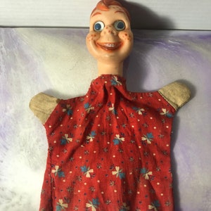 May include: Vintage hand puppet with a red and white floral dress, a smiling face with large blue eyes, and red hair.