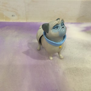 Puede incluir: Un pequeño juguete de perro pug gris y blanco con collar azul y botón amarillo. El juguete tiene una expresión de dibujos animados con ojos grandes y la boca ligeramente abierta.