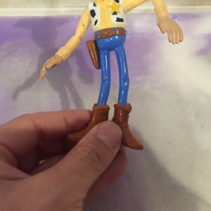 May include: A plastic toy figure of Woody from the Toy Story franchise. Woody is wearing a brown cowboy hat, a yellow vest with a red bandana, blue jeans, and brown boots. He has a sheriff's star on his vest.