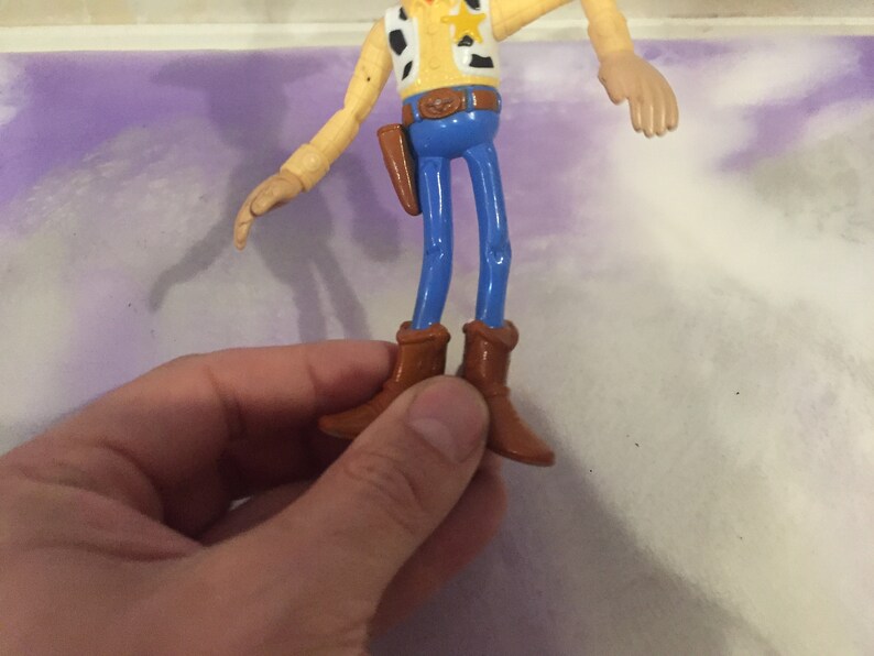 May include: A plastic toy figure of Woody from the Toy Story franchise. Woody is wearing a brown cowboy hat, a yellow vest with a red bandana, blue jeans, and brown boots. He has a sheriff's star on his vest.
