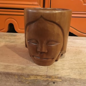 May include: A wooden cup carved with a face. The cup is brown and has a smooth finish.