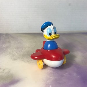 May include: A vintage Donald Duck toy in a red, white, and blue airplane. The toy features Donald Duck's iconic blue hat, yellow beak, and blue shirt. The airplane has yellow wheels and a white undercarriage.