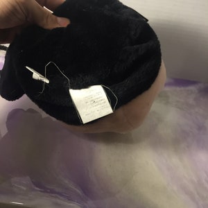 Puede incluir: Juguete de peluche negro con una etiqueta blanca adjunta. La etiqueta tiene texto que dice "Hecho en China, Material totalmente nuevo, Fibra de poli&eacute;ster, Contenido: 100% poli&eacute;ster, Cuidado: Lavar solo a mano, No blanquear, No planchar, No limpiar en seco, No secar en secadora, Hecho exclusivamente para:  " y luego el texto se corta.