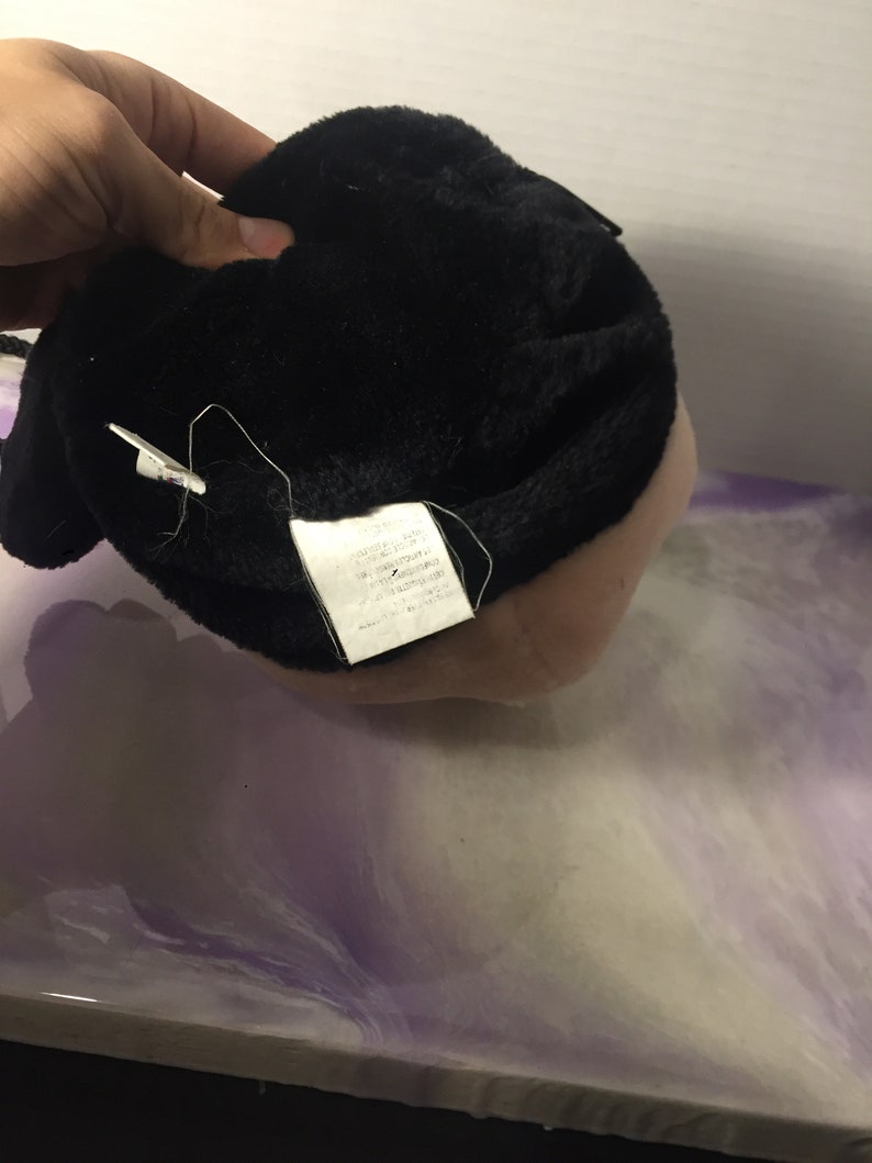 Puede incluir: Juguete de peluche negro con una etiqueta blanca adjunta. La etiqueta tiene texto que dice "Hecho en China, Material totalmente nuevo, Fibra de poli&eacute;ster, Contenido: 100% poli&eacute;ster, Cuidado: Lavar solo a mano, No blanquear, No planchar, No limpiar en seco, No secar en secadora, Hecho exclusivamente para:  " y luego el texto se corta.