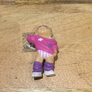 May include: A small plastic doll wearing a pink and white striped shirt with a white star, purple leg warmers, and white shoes.