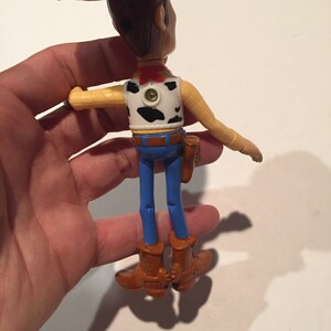 May include: A Woody action figure, viewed from the back, with a brown cowboy hat, vest with black cow spots, blue jeans, and brown boots. The figure has a red neckerchief and a holster. The arms are outstretched.