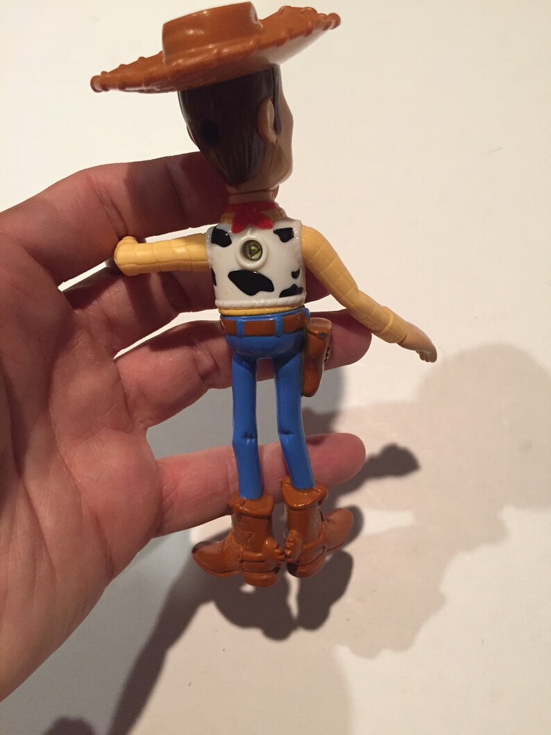 May include: A Woody action figure, viewed from the back, with a brown cowboy hat, vest with black cow spots, blue jeans, and brown boots. The figure has a red neckerchief and a holster. The arms are outstretched.