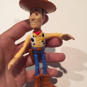 May include: A Woody action figure from the Toy Story franchise. The figure is wearing a brown cowboy hat, yellow shirt, blue jeans, and brown boots. It has a sheriff's badge and a holster. The figure is held in a hand.