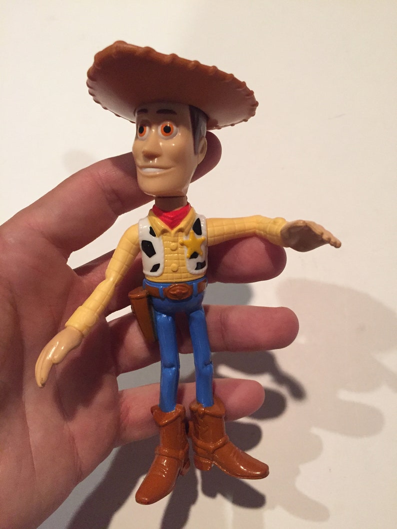 May include: A Woody action figure from the Toy Story franchise. The figure is wearing a brown cowboy hat, yellow shirt, blue jeans, and brown boots. It has a sheriff's badge and a holster. The figure is held in a hand.