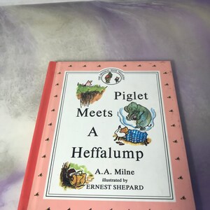 May include: A pink book with a title that reads "Piglet Meets a Heffalump" by A.A. Milne, illustrated by Ernest Shepard. The book cover features illustrations of Piglet, a Heffalump, and Winnie the Pooh.