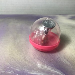 May include: A small pink and clear plastic toy capsule with a miniature figure inside. The figure is wearing a black hat and a white shirt.